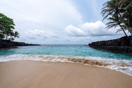 A secluded sandy beach cove framed by dark volcanic rock formations and leaning palm trees on Ilh&eacute;u das Rolas (Equator Island), S&atilde;o Tom&eacute; and Pr&iacute;ncipe