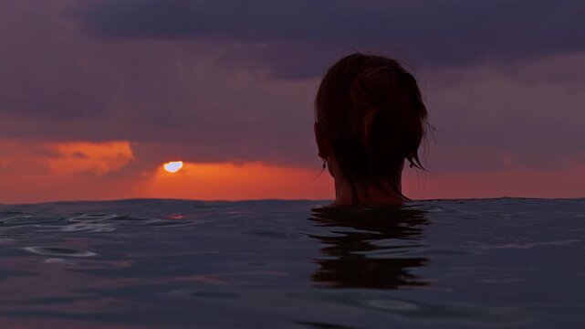 Woman floating in calm sea facing glowing sunset, back view at water level with dramatic dark cloud and orange horizon, slow motion peaceful ocean mood