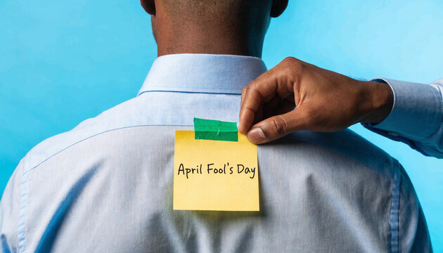 Close-up of hand attaching an april fool's day sticky note on back of man's shirt, office prank, blue background