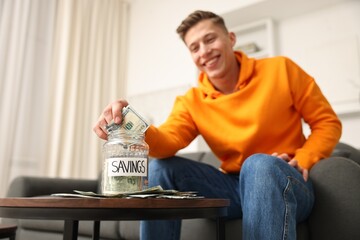 Happy man with savings jar and banknotes at wooden table indoors, selective focus