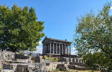 Fototapeta premium Ancient stone Garni Temple with classic columns stands on a grassy hill under a blue sky. Rugged mountains rise in the distant background. Armenia