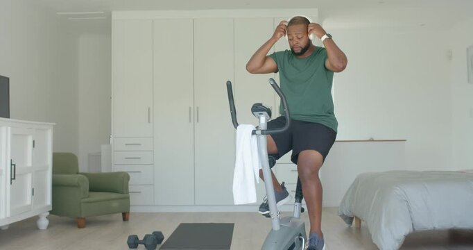 Adult African American man fitting headphones, pedaling bike in bedroom, in green tee exercising