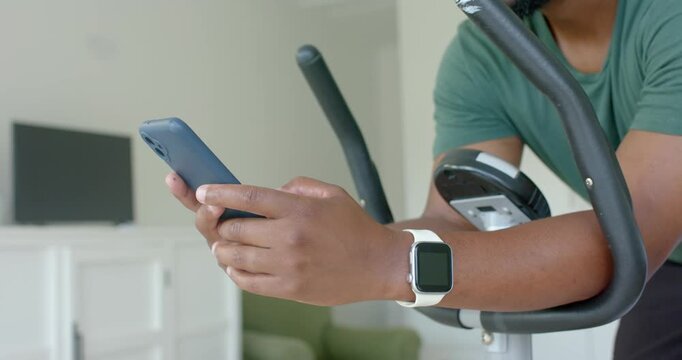 Man riding exercise bike at home scrolling phone, looking up and reacting to content, earbuds