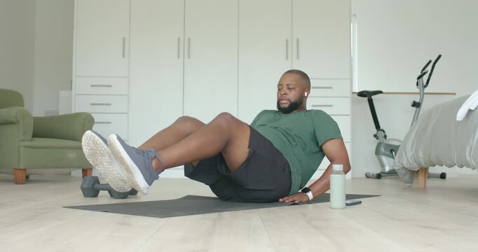 Man wearing green tee, performing seated core knee tucks on mat in bedroom, following earbuds guide