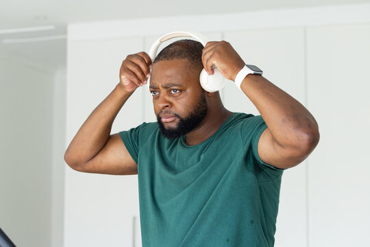 African man lifting white over-ear headphones, sporting green T-shirt and white smartwatch at home