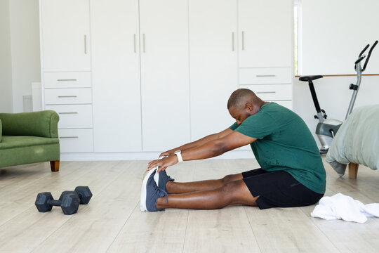African American man in green sport tee stretching on floor in bedroom, reaching toes near weights