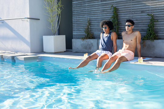 African American men sitting at pool edge with shades dipping feet and sipping drinks, copy space