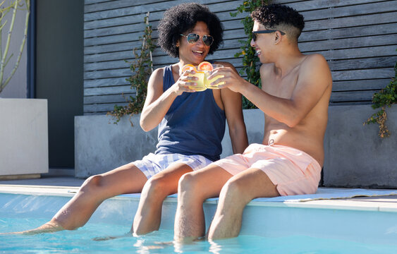 Two African men sitting on pool edge at backyard pool, in trunks, tank, clinking citrus drinks