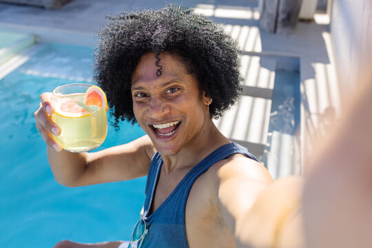 African American man raising glass and taking selfie poolside on wooden deck wearing blue tank top