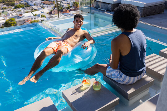 African American male friends relaxing rooftop pool, one on blue ring in orange trunks, two drinks