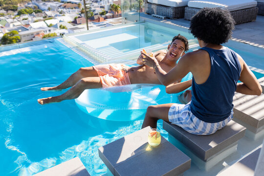 Diverse male friends reclining and reaching in swimwear on rooftop pool float, passing drink