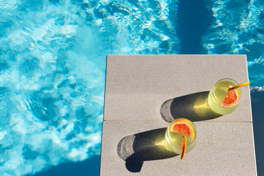 Two stemless glasses sitting on concrete pool coping, casting long shadows and rippling reflections