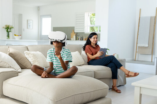 Mother and African American son sitting in living room, mother using tablet, son wearing VR headset