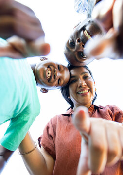 Diverse family with teen leaning in, smiling, hands reaching toward camera outdoors, bracelet seen