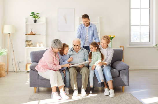Happy multi generational extended family reading a book at home. Grandparents, parents and little kids sitting on the couch in a living room interior and reading a book of children's stories together