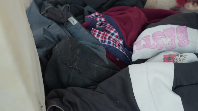 Black laundry scattered on carpet, open hamper spilling mixed garments near bedside, plaid shirt and floral beanie peeking from heap, towels and sweaters overlapped on soft rug, messy interior showing