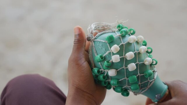 Close up black hands holding shaker. Green and white beaded net wrapped around gourd shaker, sandy fingertips and palms, slow careful inspection, textured beads and knotwork in soft natural light,