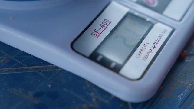 Digital kitchen scale closeup on countertop. White plastic corner on scratched blue surface, small lcd display showing faint numbers, home baker measuring ingredient for house chores and recipe prep,