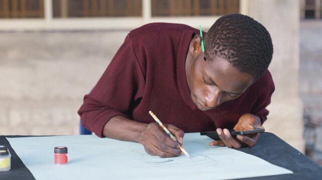 Black man painting on paper, dynamic action shot of young African artist applying ink and brush, small paint jar and smartphone reference on table, outdoor workshop vibe, intense concentration,