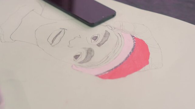 Overhead portrait sketch with phone on paper, black outlines and pink hat partially colored, smartphone resting on sheet, still studio moment, minimal composition and soft lighting, readytodocument