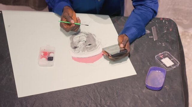 Black artist mixing black paint for demo, darkskinned male applying bold black and gray washes around pink underlay on white sheet, green brushwork, phone reference, open palette and container