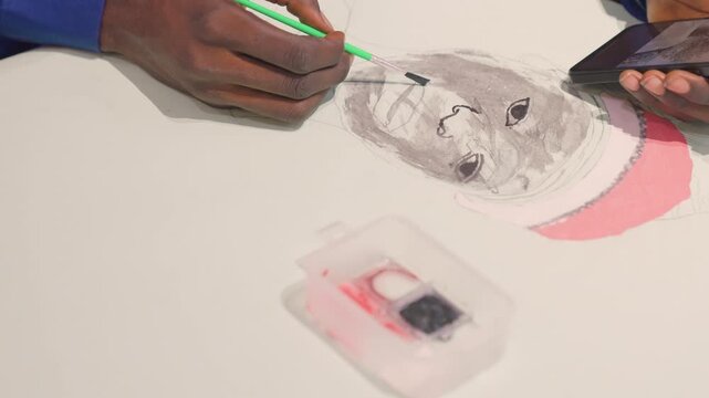 Overhead black artist adding facial details, green tool refining shading on monochrome portrait, smartphone reference held at edge, small palette box and pink hat accent, concentrated deliberate