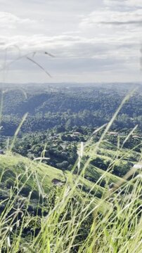 Scenic high-angle footage of lush green valleys and rolling hills in Sao Roque, Sao Paulo, Brazil. Beautiful landscape with wind blowing through grass, rural infrastructure, and a bright sky with clou