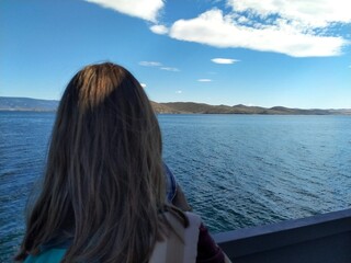 Fototapeta premium A woman looks from behind at a calm lake and distant hills under a blue sky. Travel, rest, freedom, copy space. 