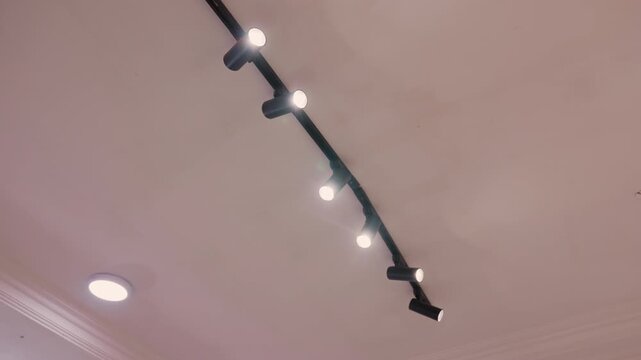 Black track lights on ceiling set modern gallery mood. Row of adjustable spotlights mounted on black track, warm led bulbs casting soft beams across plaster ceiling and decorative molding. Recessed