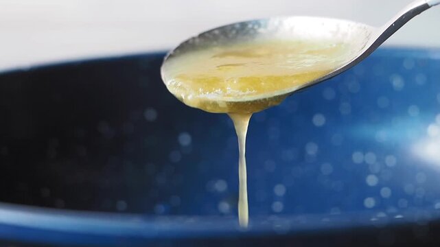 Slow motion video of liquid ghee butter dripping from silver spoon into blue cooking pan