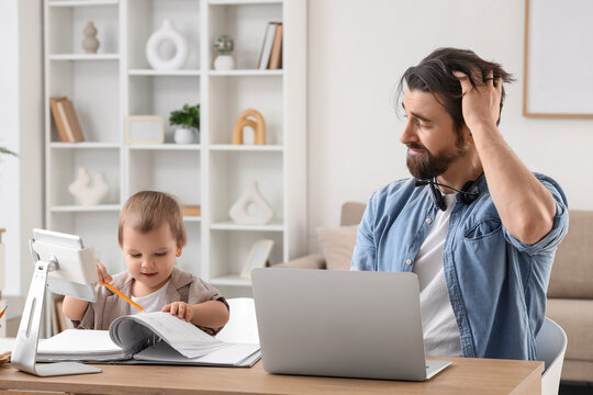 Little baby boy drawing in document with his stressed father working at desk from home during parental leave