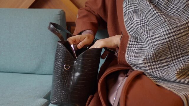 Woman searching for belongings in black leather crocodile embossed handbag sitting on blue sofa indoors
