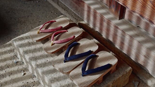 Two pairs of wooden geta sandals with red and blue straps resting on a stone step at the entrance of a historic house. Morning sunlight through a lattice door creates a beautiful striped shadow patter