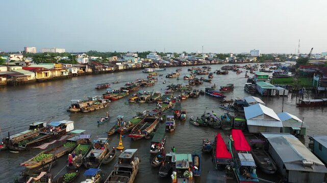 Aerial view of numerous boats trading goods at the bustling Cai Rang floating market on a river, with waterfront buildings in the background. The vibrant scene captures the energy of this iconic