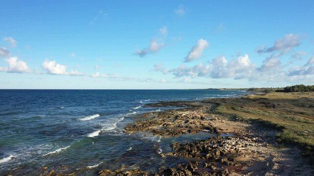 Wide view of a rugged rocky shoreline with clear blue sea and scattered clouds, captured near Brindisi, Italy.