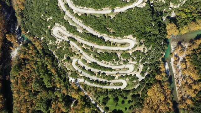 Aerial view of a serpentine mountain road cutting through lush forest in Vikos Aoos National Park, Greece. The curving road contrasts with the dense greenery and river below.