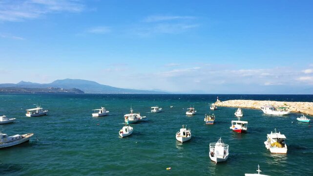 Small fishing boats float in the turquoise waters of Koroni harbor with a rocky breakwater and distant mountains under a clear blue sky. Tranquil Mediterranean coastal scene ideal for travel and