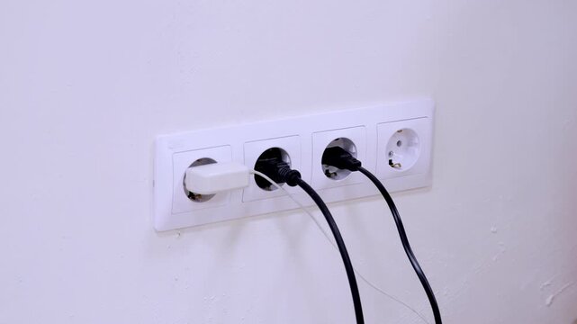 Hand plugs white plug into wall socket. Three sockets mounted on plain white wall. Black cords plugged into two adjacent sockets. Hand adjusts plug position in socket. Action captured in clear