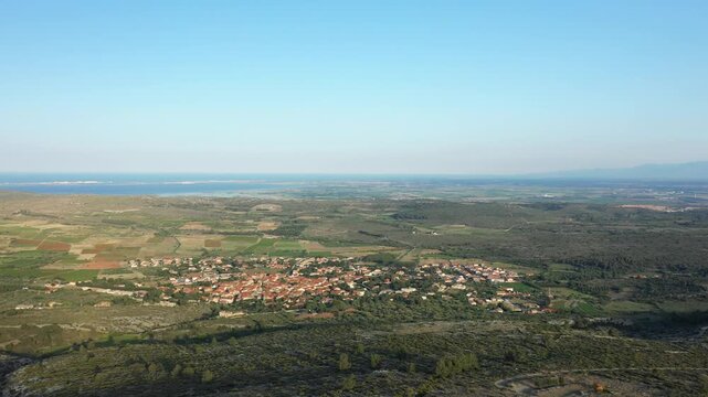 Wide aerial landscape of Opoul Perillos village and surrounding fields in southern France, with the Mediterranean Sea visible in the distance under a clear blue sky.