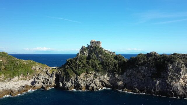 Dramatic rocky coastline with lush greenery and a historic stone tower overlooking the blue sea at Capo di Conca, Italy. Clear sky and vibrant Mediterranean landscape create a scenic view.