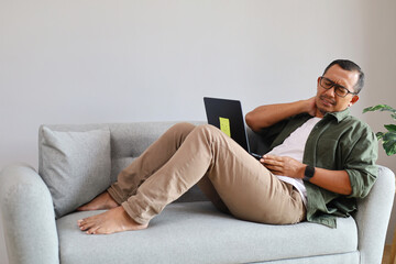 Fototapeta premium A man at home massages his sore neck while sitting on the sofa in front of his laptop, looking tired after long hours of digital work, discomfort and tension from sitting for hours and muscle tension.