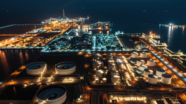 Night aerial panorama of coastal petroleum refinery complex with glowing storage tanks, pipelines, and shipping port infrastructure reflecting on dark sea, representing modern energy industry.