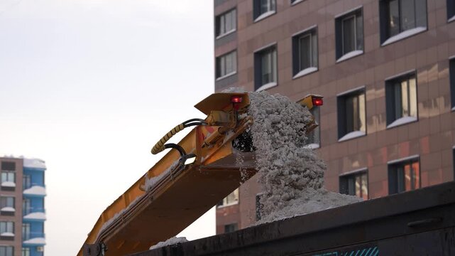 Yellow snow loader machine with a conveyor belt is loading dirty snow and ice into the back of a large dump truck body as part of the city's winter street cleaning service