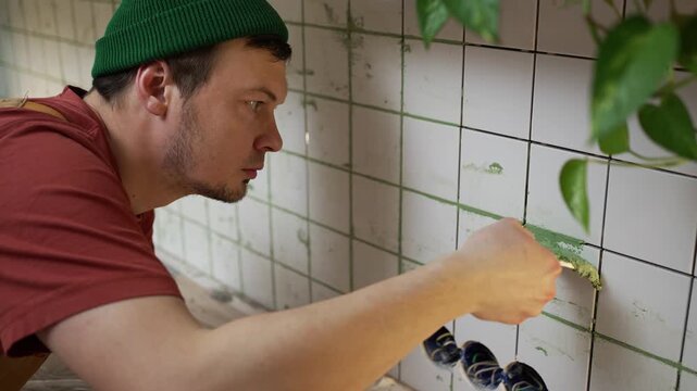 Concentrated worker using a spatula to apply green grout to the joints between white kitchen tiles. Skilled man meticulously finishing the tile installation during a home improvement project