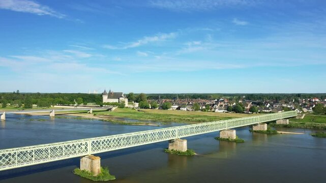 Wide view of a metal bridge crossing the Loire River with Sully sur Loire village and historic chateau in the background under a clear blue sky.
