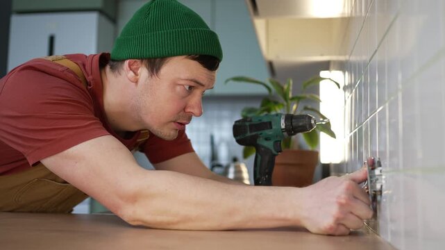 Young man in uniform installing a new power outlet on a kitchen apron, a skilled electrician using a cordless drill to mount the socket during home improvement or repair