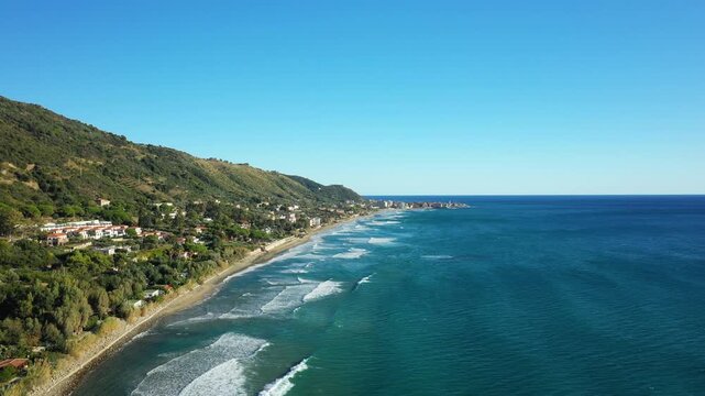 Scenic aerial view of the coastline and turquoise sea in Acciaroli, Italy, with waves gently rolling onto the sandy beach and lush green hills in the background.