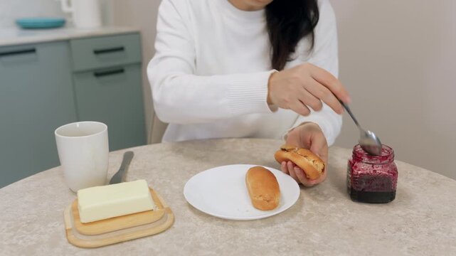 A woman in a white sweater sits at a kitchen table preparing a snack. She holds a bun and uses a spoon to scoop jam from a jar to spread on it. Butter and a mug are on the table.