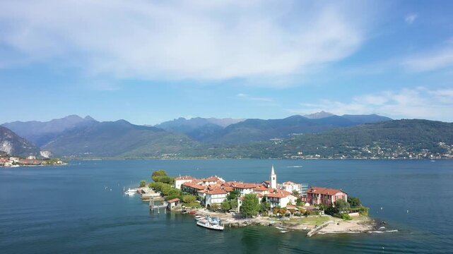 Aerial view of Isola dei Pescatori, a picturesque island with traditional buildings and a church tower on Lake Maggiore, Italy, surrounded by mountains and blue water under a clear sky.