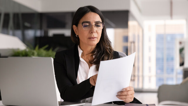 Aged professional woman sits at workplace desk with open laptop in modern office, holding papers, analyzing document or reviewing financial report, thinking on received information or important data